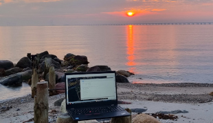 Fotografi af en laptop i strandkanten ved solopgang.